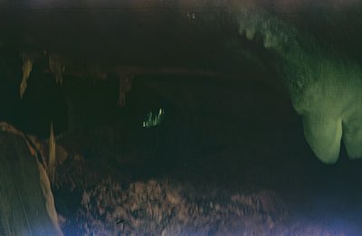 A wide, dark cave interior. Limestone formations, likely stalactites and stalagmites, define the cavern walls. A narrow slit ...