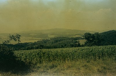 Lush, terraced cornfields dominate the midground, sloping toward a distant, hazy mountain range. Dense forest lines the hills...