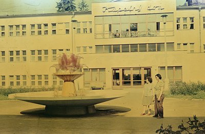 A wide, tiered fountain dominates the foreground of a vintage photograph. A modern, multi-story building with a grid of windo...