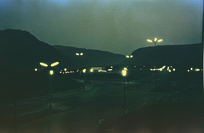 Night view of a town nestled within a mountainous landscape. Prominent, stylized lampposts illuminate a wide plaza or courtya...