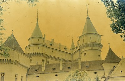 A vintage, sepia-toned photograph depicts a stately castle with prominent conical towers and crenellated battlements. Archite...