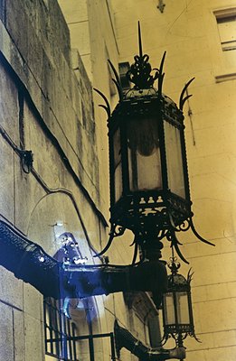 Ornate, wrought-iron chandelier hangs from a high ceiling, reflecting light within its panes. The architectural setting sugge...