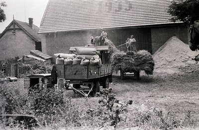 Hay harvest scene. A trailer loaded with bundled hay and sacks is parked near a farm building. Two figures are visible on the...