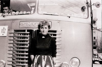 A young woman stands proudly beside a Škoda bus, likely in Czechoslovakia. She wears a dark, textured sweater and a full, pat...