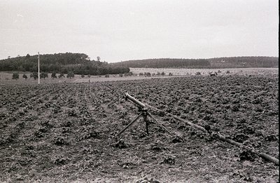 A field artillery piece, likely a World War I-era field gun, sits mounted on a tripod within a cultivated field. The surround...