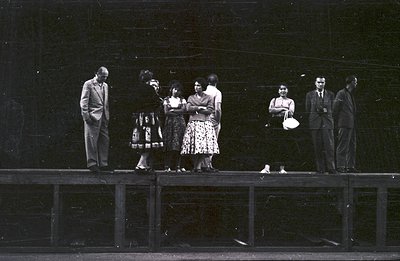 Group of seven individuals, six adults and one child, stand on a raised platform or stage, facing the camera. Their attire su...