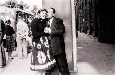 A smiling couple embraces near a building exterior, likely at a transit station. The woman wears a patterned full-skirted dre...