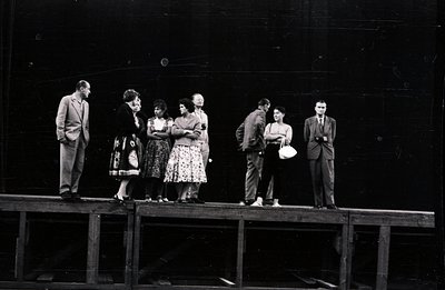 Six individuals stand on a wooden platform against a dark, possibly stage backdrop. Men in suits and a woman in a dress appea...