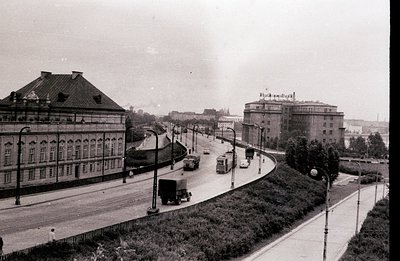 An elevated, wide avenue curves into the distance, featuring early automobile and truck traffic. A monumental, classically-st...