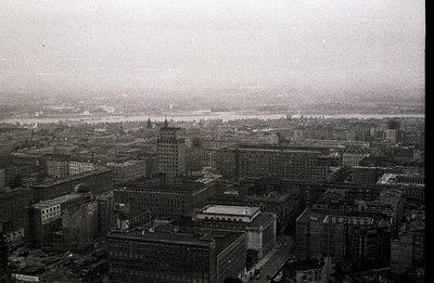 A high-angle, monochrome cityscape shows dense urban architecture along a wide river. Buildings display early 20th-century de...