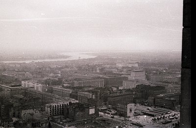 High-angle view of a cityscape featuring dense, mid-century architecture. A wide river dominates the background, partially ob...