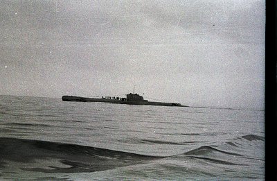 A large submarine cuts through a choppy sea. The vessel's conning tower and deck structures are visible against a heavily ove...