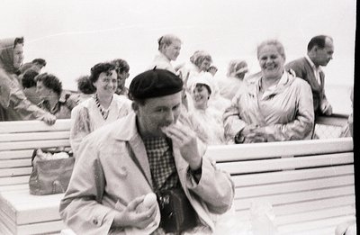 A group of people, likely tourists, are seated on a boat's deck. A man in a beret and light jacket appears central, holding a...