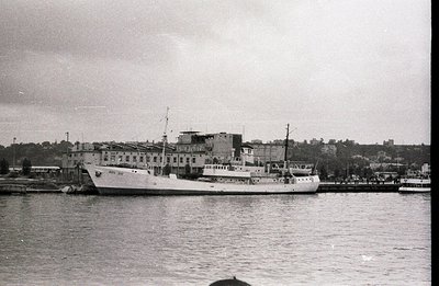 A coastal research vessel, "RV 312", is docked alongside a pier, viewed against a backdrop of a large, multi-story building, ...