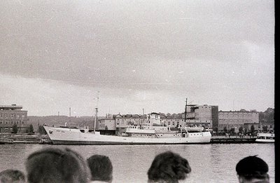 A medium-sized cargo ship is docked at a pier, framed by a multi-story industrial or administrative building. Partial view of...