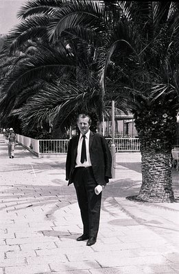 Elegant man in a dark suit and tie stands formally beside a palm tree, likely a seaside promenade. He holds a rolled document...