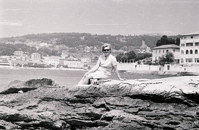 A woman in a knee-length dress and sunglasses sits casually on rocky coastline. Behind her, a seaside town with tiered buildi...