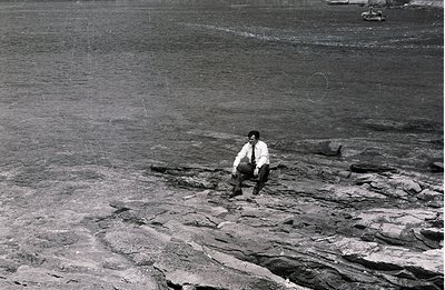 Man in a suit sits cross-legged on a rocky shoreline. The textured stone contrasts with dark water, likely a seaside or coast...
