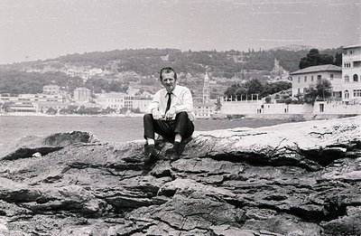 Man in a suit sits on rocks overlooking a seaside town and harbor. Appears to be a promotional portrait, perhaps for tourism ...