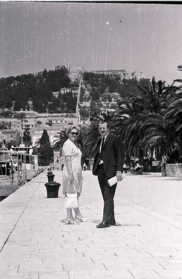 A formally dressed couple poses on a stone promenade, likely during a vacation. The woman wears a sheath dress and carries a ...