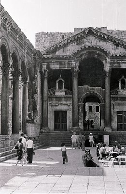 A view of a monumental Roman-era building featuring a large courtyard with arched porticoes and Corinthian columns. People ex...