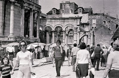 Black and white photo depicting a bustling street scene featuring ancient Roman architecture. Tourists and locals navigate a ...