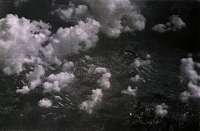 High-altitude aerial view showcases a rugged, mountainous landscape partially obscured by voluminous clouds. Visible textures...