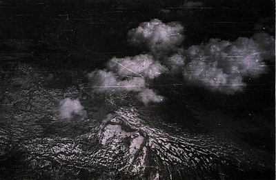 Dramatic aerial view depicts a snow-covered mountain range obscured by heavy, swirling clouds. Visible texture suggests a vin...