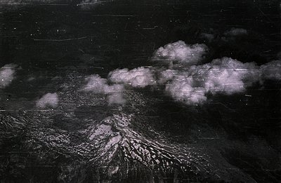 Aerial view of a snow-covered, volcanic peak, partially obscured by swirling cloud formations. The image exhibits significant...