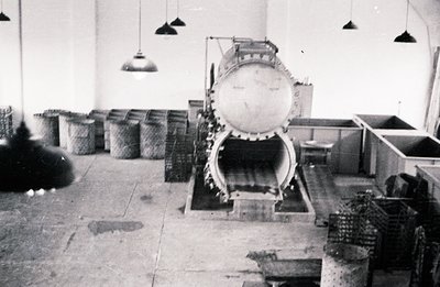 Industrial scene with a large, vintage grain processing machine dominating the space. Woven baskets line the left wall; metal...