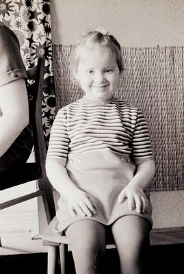A young girl sits formally in a chair, smiling broadly. She wears a striped top, high-waisted shorts, and a headband. The bac...
