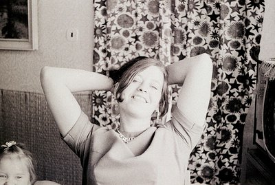 A smiling woman sits on a sofa with her arms behind her head, seemingly relaxed. Detailed floral wallpaper and a vintage tele...