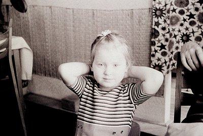 A young girl sits with her hands behind her head, displaying a direct gaze. She wears a striped top and shorts, seated in a w...