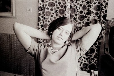 Black and white portrait of a young woman in a V-neck top & statement jewelry, hands behind her head. Likely 1960s/70s fashio...