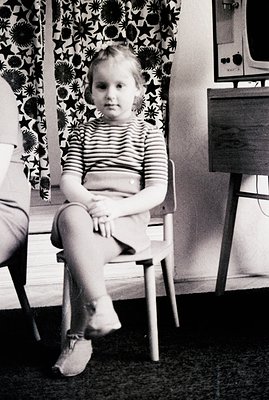 A young girl sits on a wooden chair in a domestic setting. She wears a striped shirt and shorts. A patterned curtain and a mi...