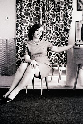 Elegant portrait of a woman in a short-sleeved dress and pearl necklace, seated on a chair with legs extended. The backdrop f...