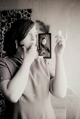 A woman examines her face in a handheld mirror. She wears a sleeveless dress, dangling earrings, and a necklace. A framed bot...
