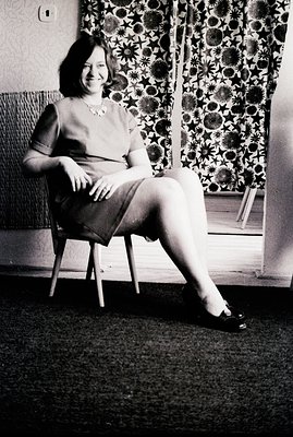 A woman sits in a wooden chair, posed for a studio portrait. She wears a fitted dress, pearl necklace, and dark shoes. Backgr...