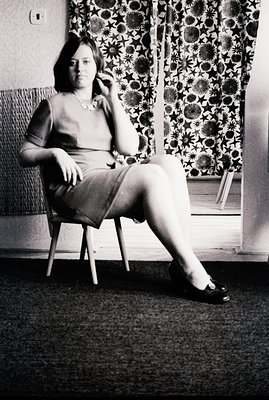 Black and white portrait captures a woman seated in a chair, exhibiting a poised yet contemplative expression. She wears a fi...