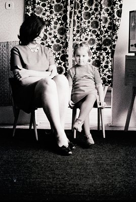 A woman and young child sit on chairs in front of patterned curtains. The woman wears a dress and heels; the child is in a st...