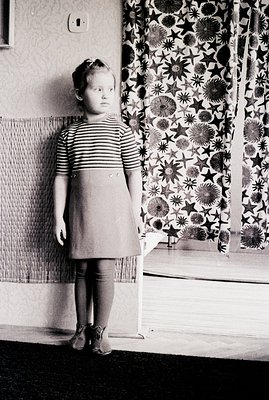 A young girl stands indoors, wearing a striped top and A-line skirt. The backdrop features patterned curtains and textured wa...
