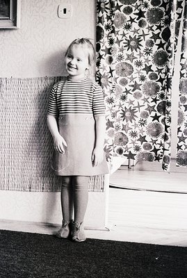 A young girl stands in a domestic interior, wearing a striped top and a simple A-line dress. She smiles directly at the camer...