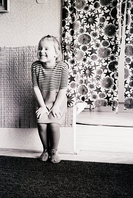 A young girl, likely 5-7 years old, sits on a textured rug, beaming at the camera. She wears a striped top, a dark skirt, and...