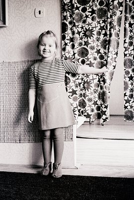 A young girl in a striped shirt and A-line dress stands with a hand on a patterned curtain. Interior scene; likely a home set...