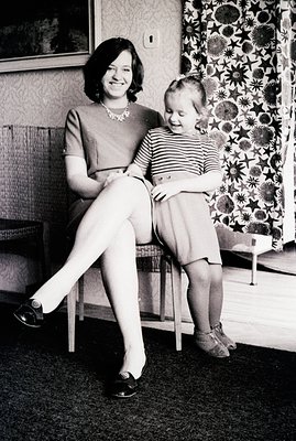 A woman and young girl pose in a home interior. The woman, seated, wears a dark dress and loafers; the girl, in a striped top...