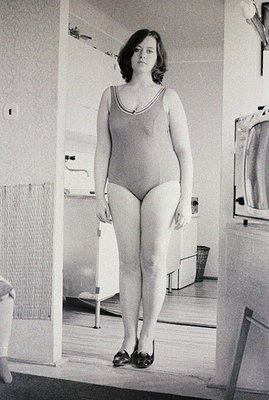 Full-length portrait of a young woman in a one-piece swimsuit, posed in a stark, institutional bathroom. The setting includes...