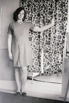 Young woman stands poised in a button-front dress, elevated platform shoes, and pearl necklace. Interior studio portrait with...