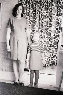 A young woman and child stand before a patterned wall. The woman wears a buttoned dress with a shallow neckline, accessorized...