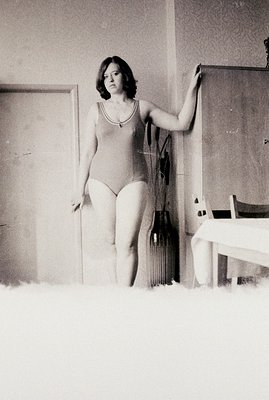 A young woman in a tank leotard poses indoors, leaning against a doorframe. The room appears sparsely furnished with a draped...