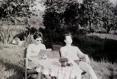 Two figures relax outdoors in a garden setting. One, seated and reading, wears a short-sleeved shirt and dark shorts. The oth...